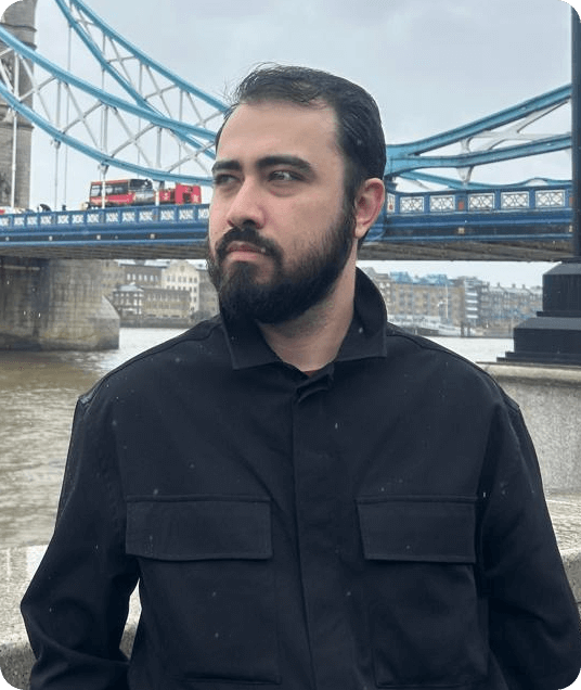 Foto do Bruno Silva, homem com barba e cabelo escuro olhando para o lado, vestindo uma jaqueta preta, em frente à Tower Bridge, em Londres, em um dia nublado.